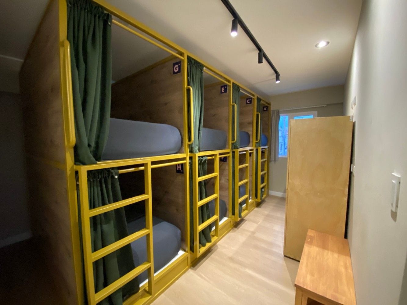 Een rij stapelbedden met gordijnen, een houten bankje en een deur.