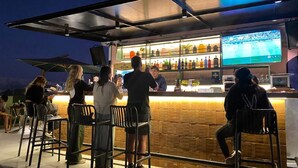 Un área de bar al aire libre con mostrador, taburetes y una pantalla mostrando un evento deportivo.