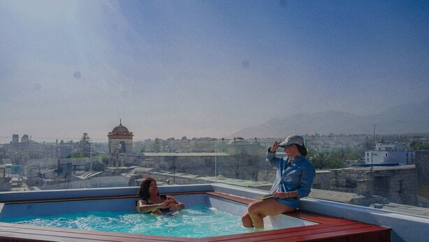Una piscina en la azotea con dos personas relajándose. La piscina está rodeada por una terraza de madera. En el fondo, se observa un paisaje urbano con edificios y una estructura en forma de cúpula.