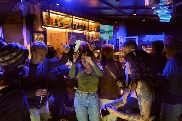 Un bar con un letrero de neón, estantes con botellas y gente conversando.