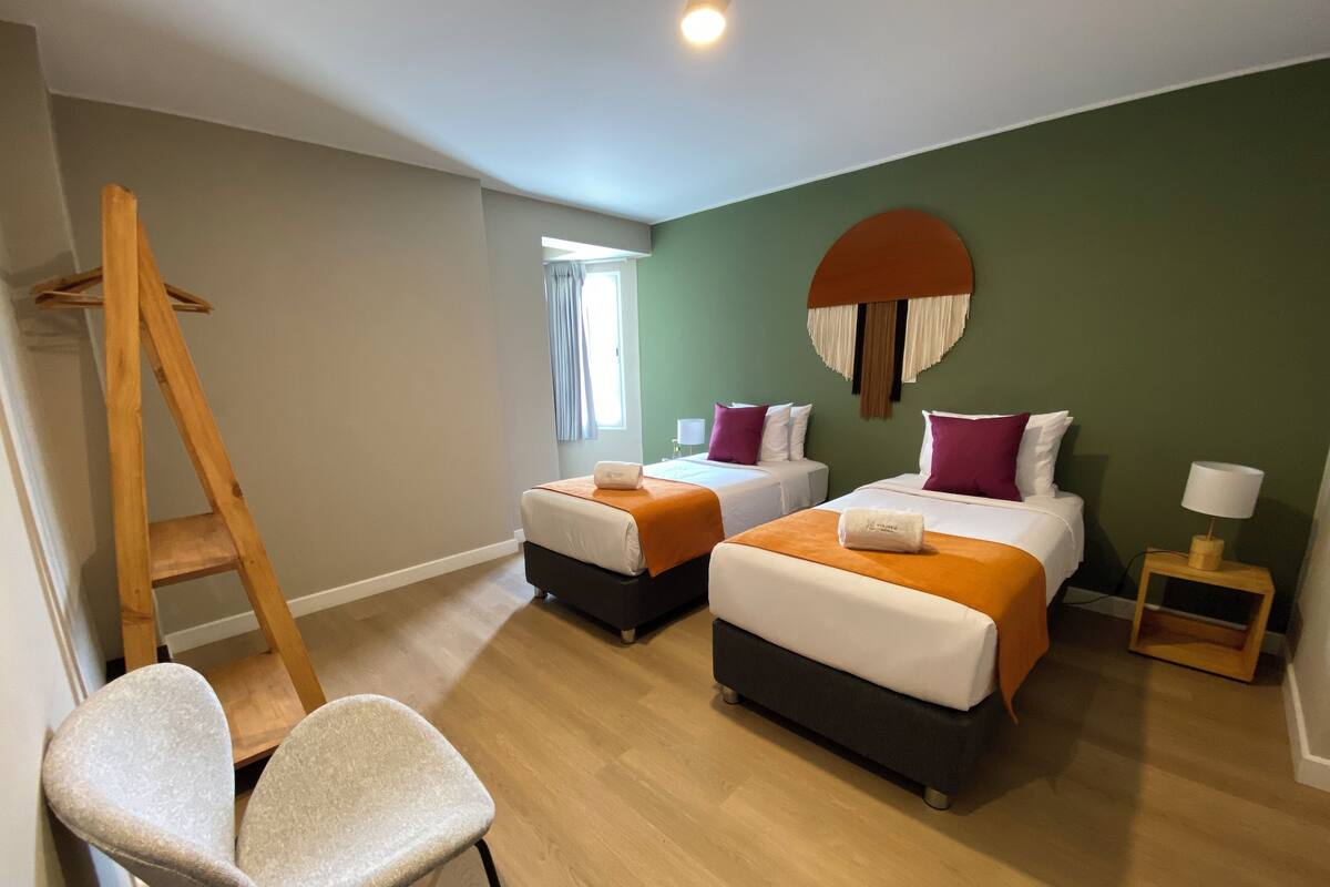 A hotel room with two beds, a wooden ladder, a chair, and a small table with a lamp.