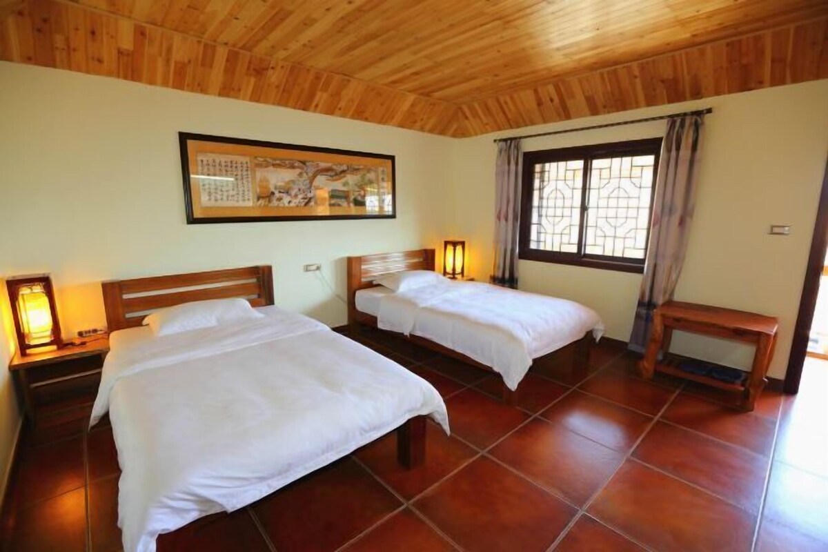 A room with two beds, wooden headboards, a nightstand with a lamp, a window with curtains, and a framed picture on the wall.