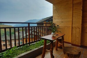 Een houten balkon met een tafel, stoelen, een bloempot en een uitzicht op verre gebouwen en bergen.