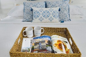 Una bandeja de mimbre con dos tazas blancas, una revista titulada 'Charleston' y una revista de 'Food & Wine' sobre una cama con ropa blanca y almohadas estampadas.