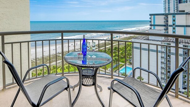 A balcony with a glass table and chairs, overlooking a beach and a pool. - Stylish Escape Oceanview Suite @ Meridian Plaza! (Myrtle Beach)