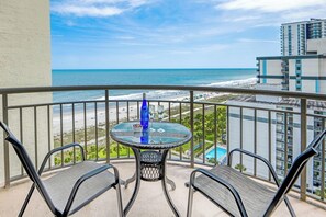 A balcony with a glass table and chairs, overlooking a beach and a pool.