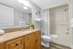 A bathroom with a granite countertop sink, wooden cabinets, a white toilet, and a glass-enclosed shower. - Luxurious Haven, Oceanview Suite (Myrtle Beach)