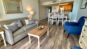 A living room with a sofa, coffee table, and blue armchair. There is a dining area with a table and chairs. - Luxurious Haven, Oceanview Suite (Myrtle Beach)
