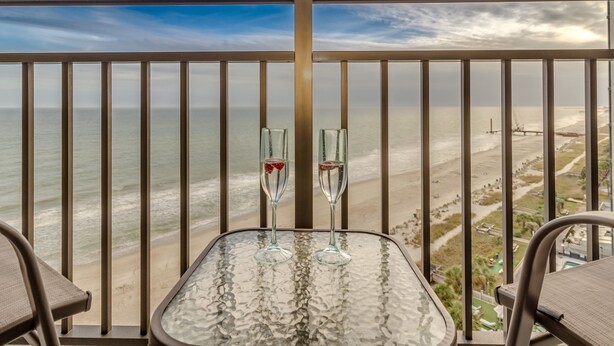 A balcony with a glass table and two champagne flutes, overlooking a beach and ocean. - Stunning Oceanfront Studio Suite! (Myrtle Beach, SC 29577)