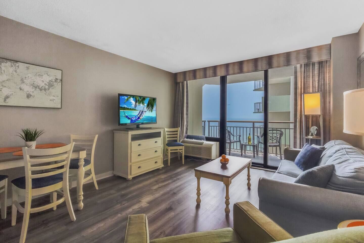 A modern hotel room with a balcony, a dining area, a sofa, and a TV on a cabinet.