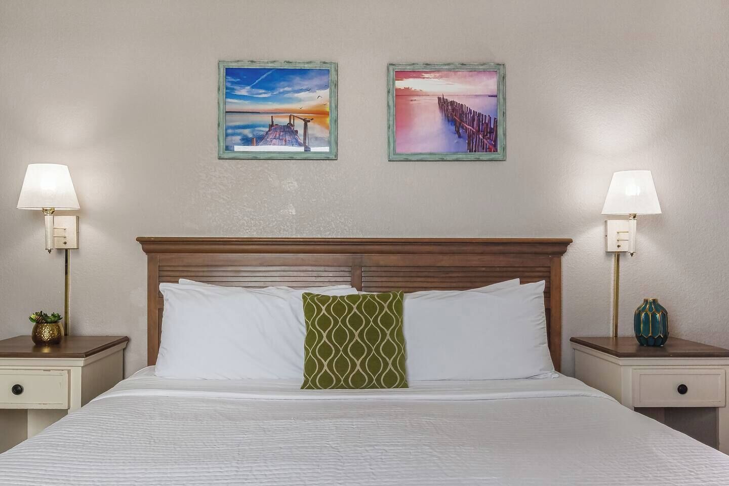 Een hotelkamer met een houten hoofdeinde, een bed met witte en groene kussens, twee nachtkastjes met lampen en twee ingelijste foto's aan de muur.
