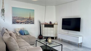 A living room with a beige sofa, a glass coffee table, a TV on a white stand, a framed cityscape picture, and a decorative vase. - Townhouse Lotus in a prime location! 5 minutes from Legoland. families & pets. (Günzburg)
