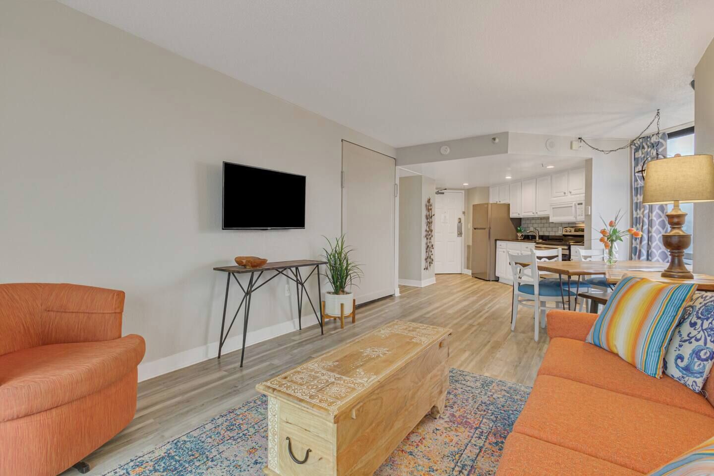 A modern living room with a sofa, a coffee table, and a TV. There is a kitchen area in the background with a fridge and a dining table.