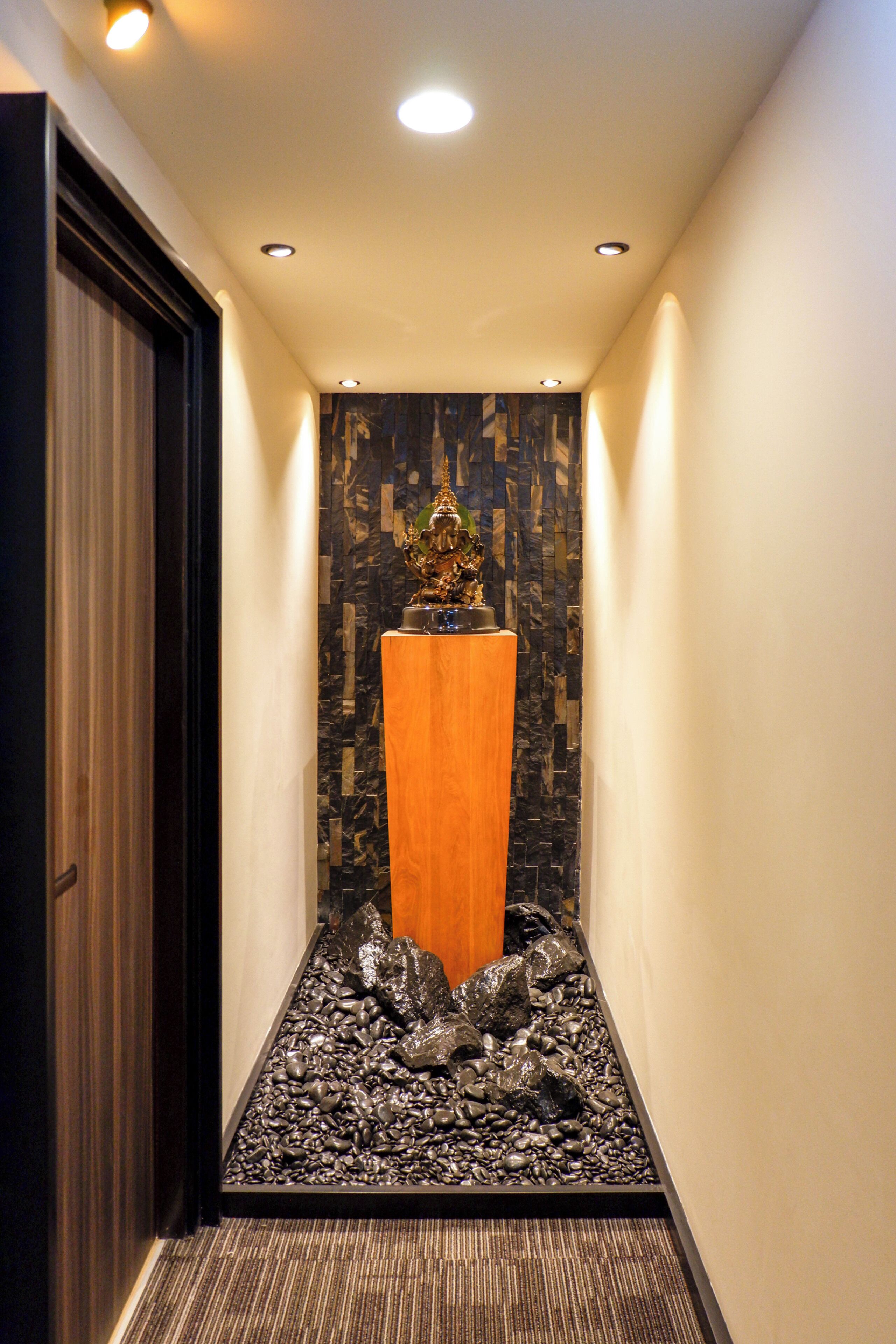 A narrow hallway with a wooden pedestal in the center, surrounded by a patterned wall and a gravel path.