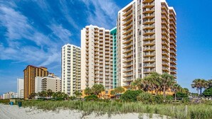 A beachfront hotel with multiple high-rise buildings, featuring balconies and a sandy beach in the foreground.