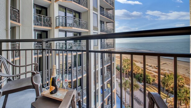A balcony with a view of the beach, a bottle of champagne, and a glass on a table.