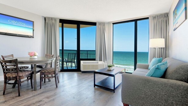 Une chambre d’hôtel avec un balcon, un coin repas avec des chaises et une table, un canapé et un téléviseur diffusant l’image d’un « RESORT CÔTIER ».