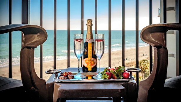 A balcony with a table set for two, featuring a bottle of champagne, two glasses, a tray with snacks, and a view of the beach. - Grand Oceanfront 1Br Suite! Dogs OK! (Myrtle Beach)