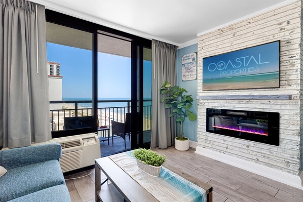 A modern living room with a large window, a fireplace, and a television displaying 'Coastal Properties'. - Inviting 1BR Oceanview Suite/Dogs OK (Myrtle Beach)