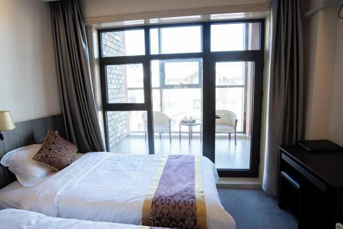 A hotel room with a large window, a bed with a patterned bedspread, a desk, and a chair visible through the window.