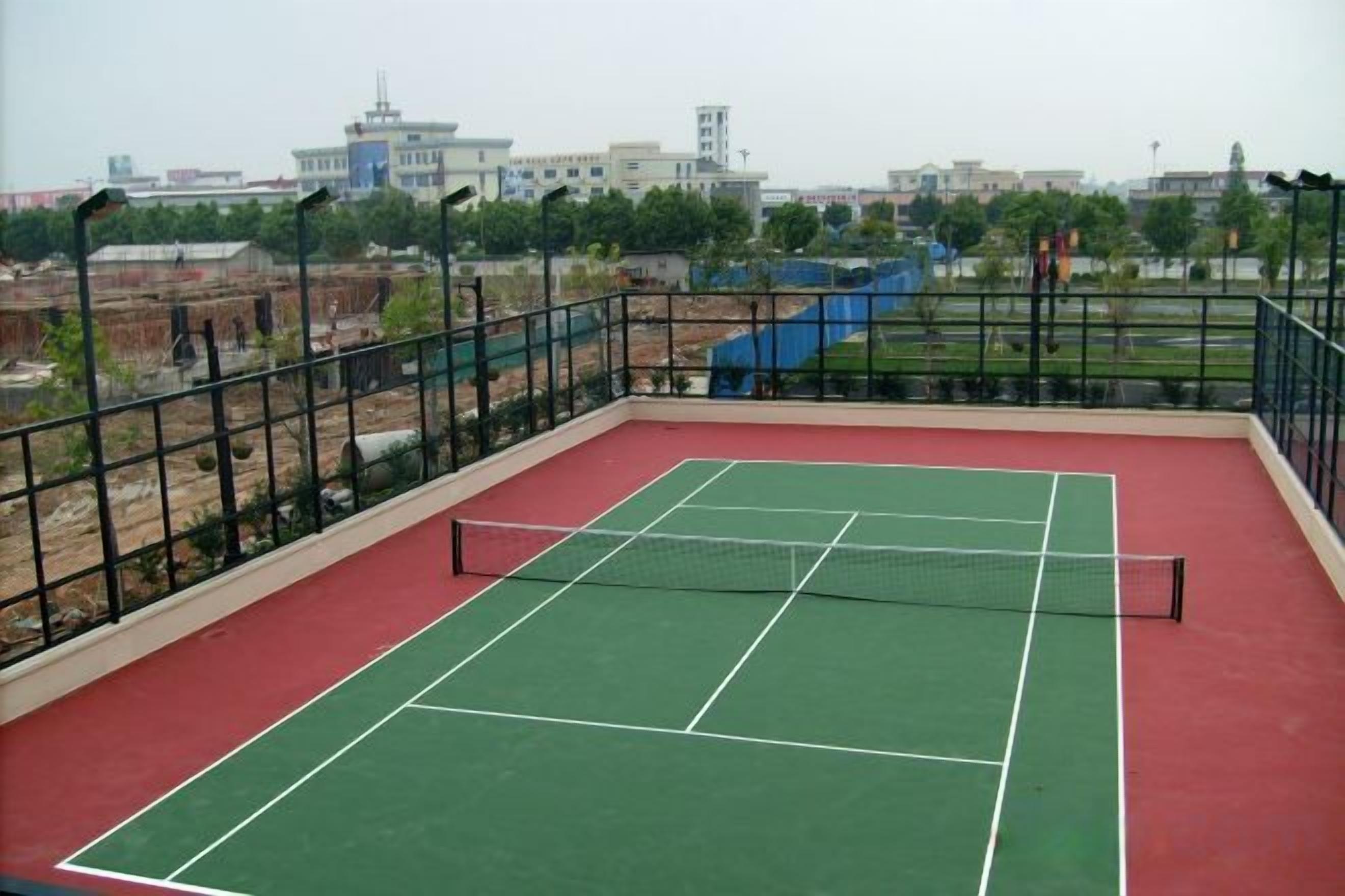 An outdoor tennis court with a net, surrounded by a metal fence, and a view of buildings in the background.