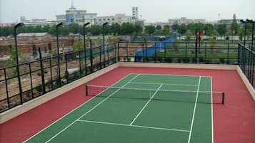 An outdoor tennis court with a net, surrounded by a metal fence, and a view of buildings in the background.