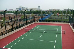 Un court de tennis en plein air, doté d’un filet et entouré d’une clôture métallique, avec en arrière-plan une vue sur des bâtiments.