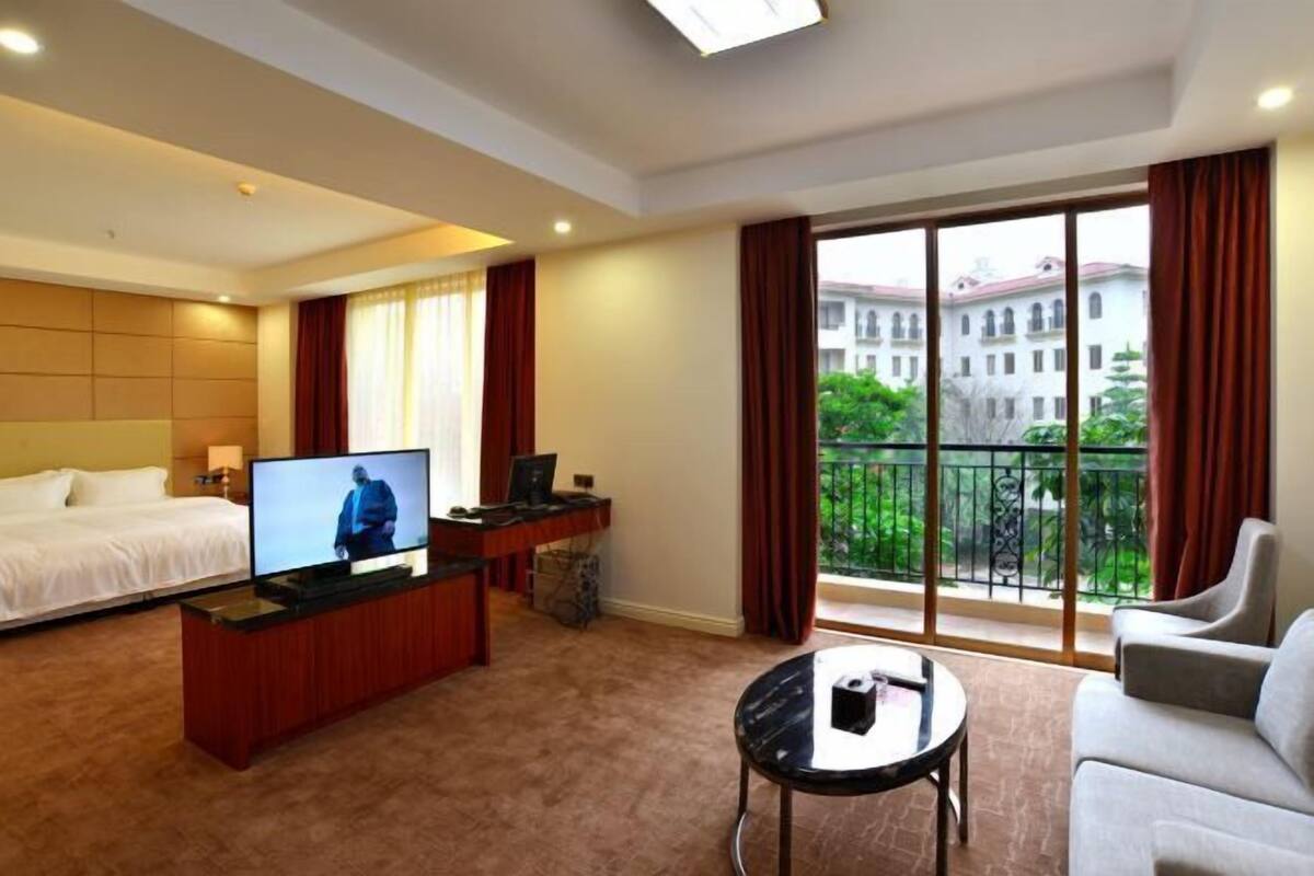 Habitació d'hotel amb llit, televisió, escriptori, sofà i balcó amb vista a un edifici i vegetació.