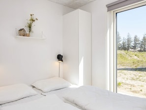A bedroom with a bed, pillows, a nightstand, a wall-mounted lamp, a shelf with a house model and flowers, and a window with a view of trees and a beach. - 5 Star Holiday Home in Romo (Rømø)