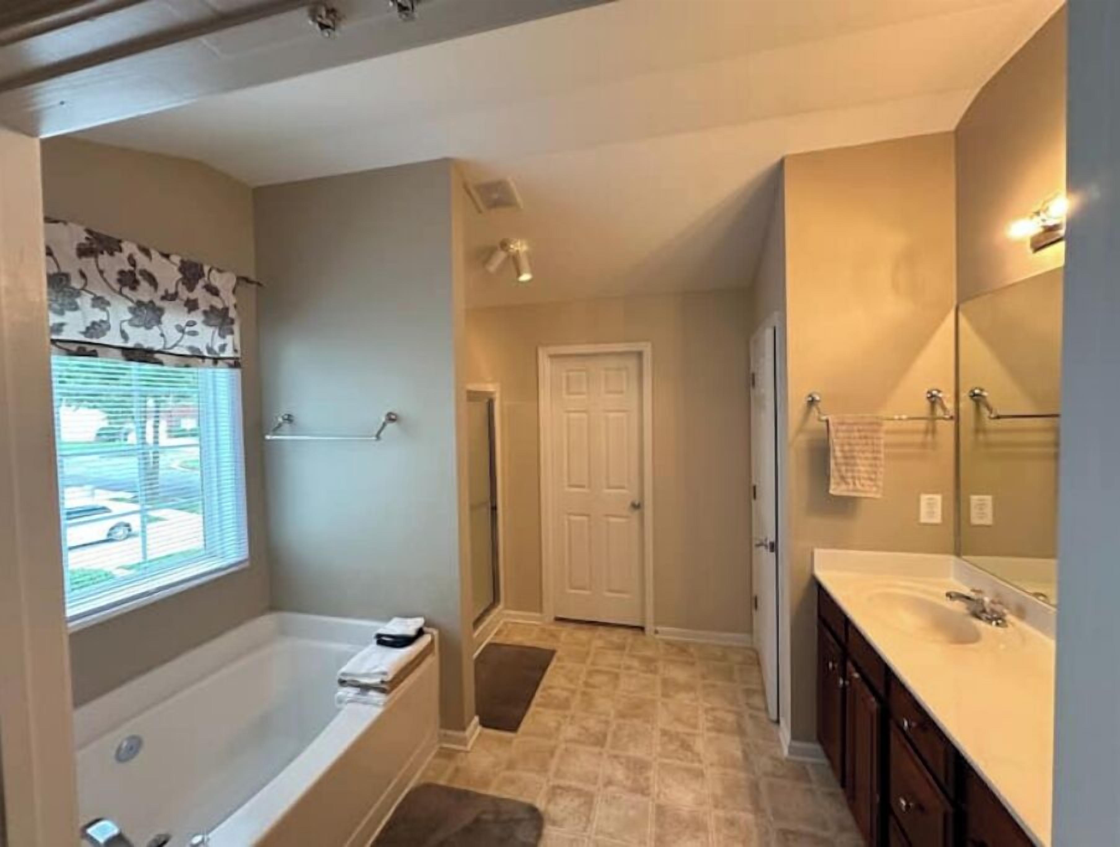 A bathroom with a bathtub, a sink, and a mirror. There is a window with blinds and a door leading to another room.