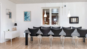 Un espace de salle à manger moderne avec une longue table et des chaises noires, une console blanche et une fenêtre donnant sur un mur de briques et des plantes.