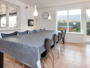 Uma sala de jantar moderna com uma mesa comprida, cadeiras pretas, luminárias pendentes, um relógio na parede e janelas amplas com vista para o exterior.