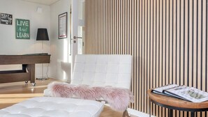 Une chambre moderne avec une tête de lit blanche à motifs capitonnés, un pouf rose moelleux, un plancher en bois, un mur à rayures et une petite table ronde sur laquelle repose un magazine.
