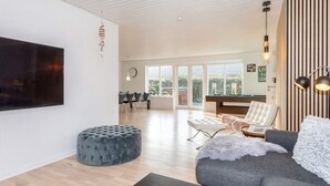 Un salon moderne avec un téléviseur à écran plat, un pouf capitonné, un canapé gris agrémenté d’un plaid moelleux, et une table de billard en arrière-plan.
