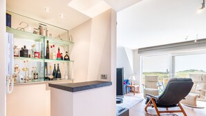 Un área de bar moderna con estantes de vidrio que exhiben varias botellas, una zona de estar con una silla negra y un sofá, y vistas al exterior a través de un ventanal grande.