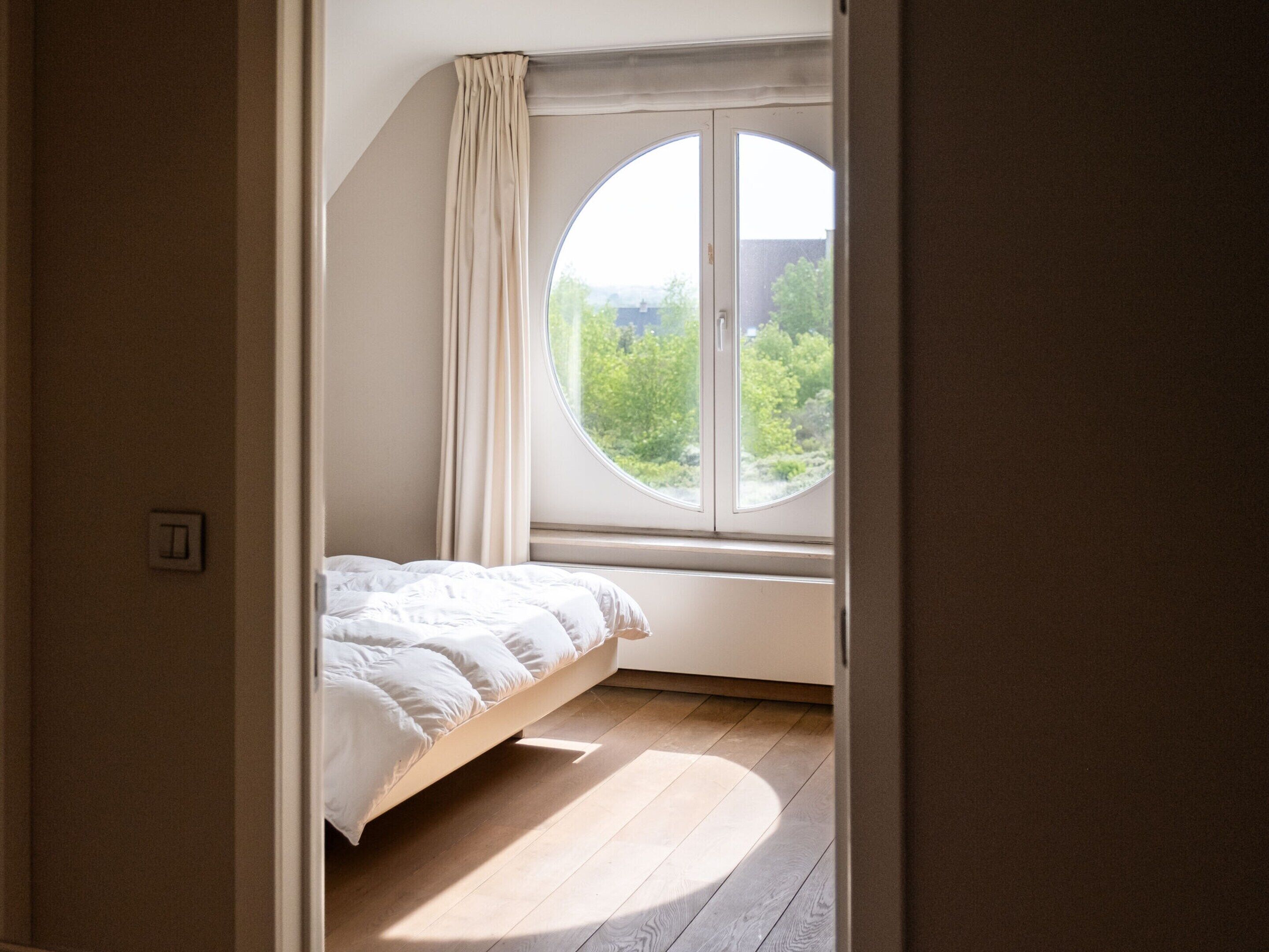 A bedroom with a bed, a round window, and a view of greenery outside.