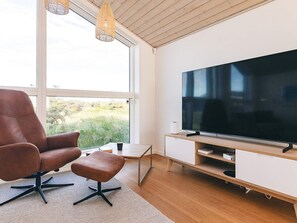 A modern living room with a large flat-screen TV, a brown leather chair, a small table, and a large window offering a view of greenery.