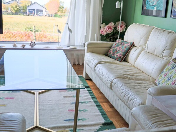 Un salon avec une table basse en verre, un canapé agrémenté de coussins à motifs, un tapis à fleurs et une vue sur une cour clôturée.