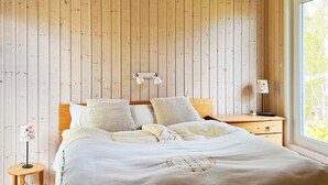 Un dormitorio con pared de listones de madera, cama con ropa de cama blanca, mesita de noche con lámpara y ventana con vistas a zonas verdes.