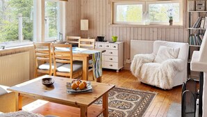Interior acogedor de una cabaña con paredes de madera, un comedor con mesa y sillas, un sillón blanco mullido y una estantería.