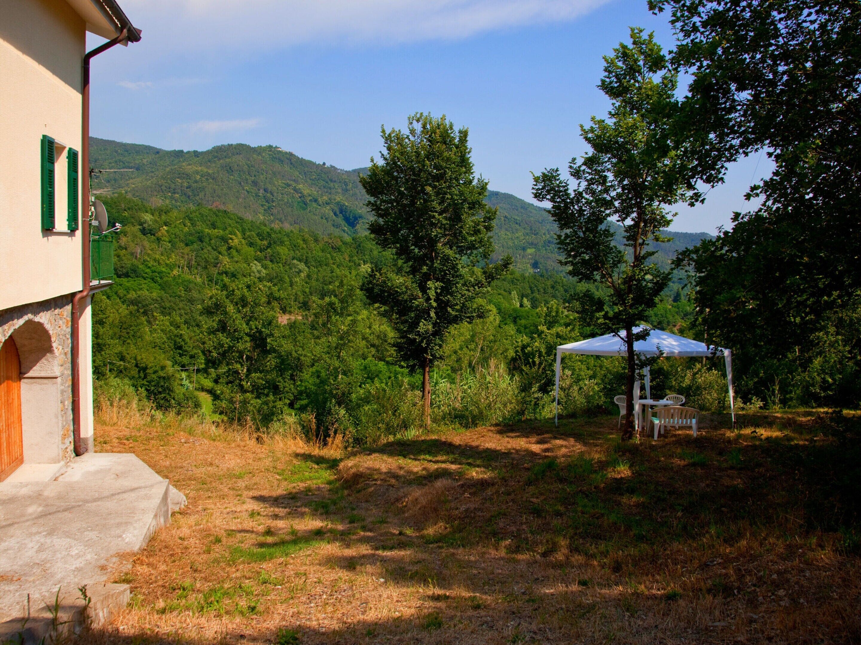 A rustic building with a white exterior and green shutters, a concrete staircase, and a white tent with chairs in a grassy area with trees and hills in the background.
