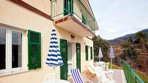 A balcony with white chairs and blue-striped umbrellas, a white table, and a building with green shutters and a balcony railing. - Cottage in Sesta Godano Near the Sea (Sesta Godano)