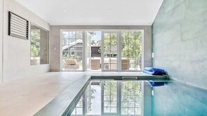 Une piscine intérieure moderne, dotée d’une paroi en verre, de grandes portes-fenêtres coulissantes donnant sur un patio extérieur, et d’un aménagement intérieur épuré et minimaliste.