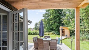 Une terrasse en bois, agrémentée de meubles en osier et d’une table, le tout entouré de verdure.