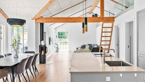 Une cuisine moderne dotée d’un îlot central, de poutres en bois, de grandes fenêtres et d’un coin repas avec une table et des chaises.