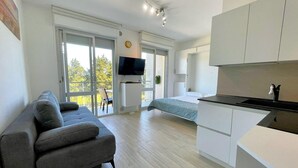A modern apartment with a grey sofa, a bed, a TV, and a balcony with a view of trees. - Apartment Porto Santa Margherita by Beach (Caorle)
