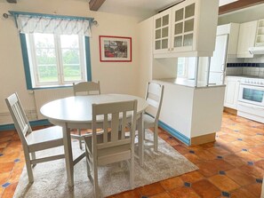 A dining area with a round table and four chairs, a window with curtains, a framed picture on the wall, and a kitchen with white cabinets and a stove. - 4 Person Holiday Home in Lovestad (Lövestad)