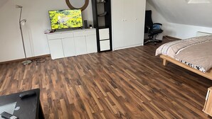 Ett sovrum med trägolv, en säng, ett skrivbord med en dator, en stol, en tv på ett stativ och en garderob.