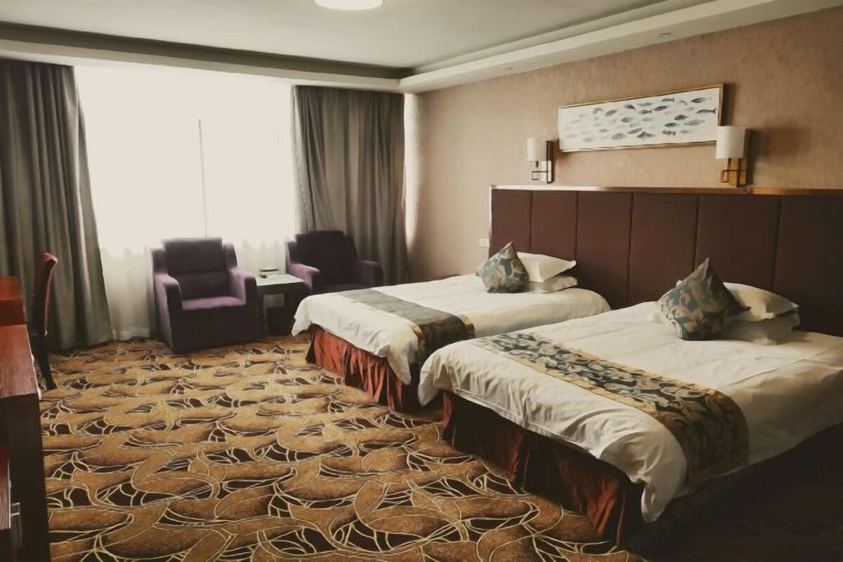 Quarto de hotel com duas camas, uma escrivaninha, uma cadeira e uma janela grande com cortinas.
