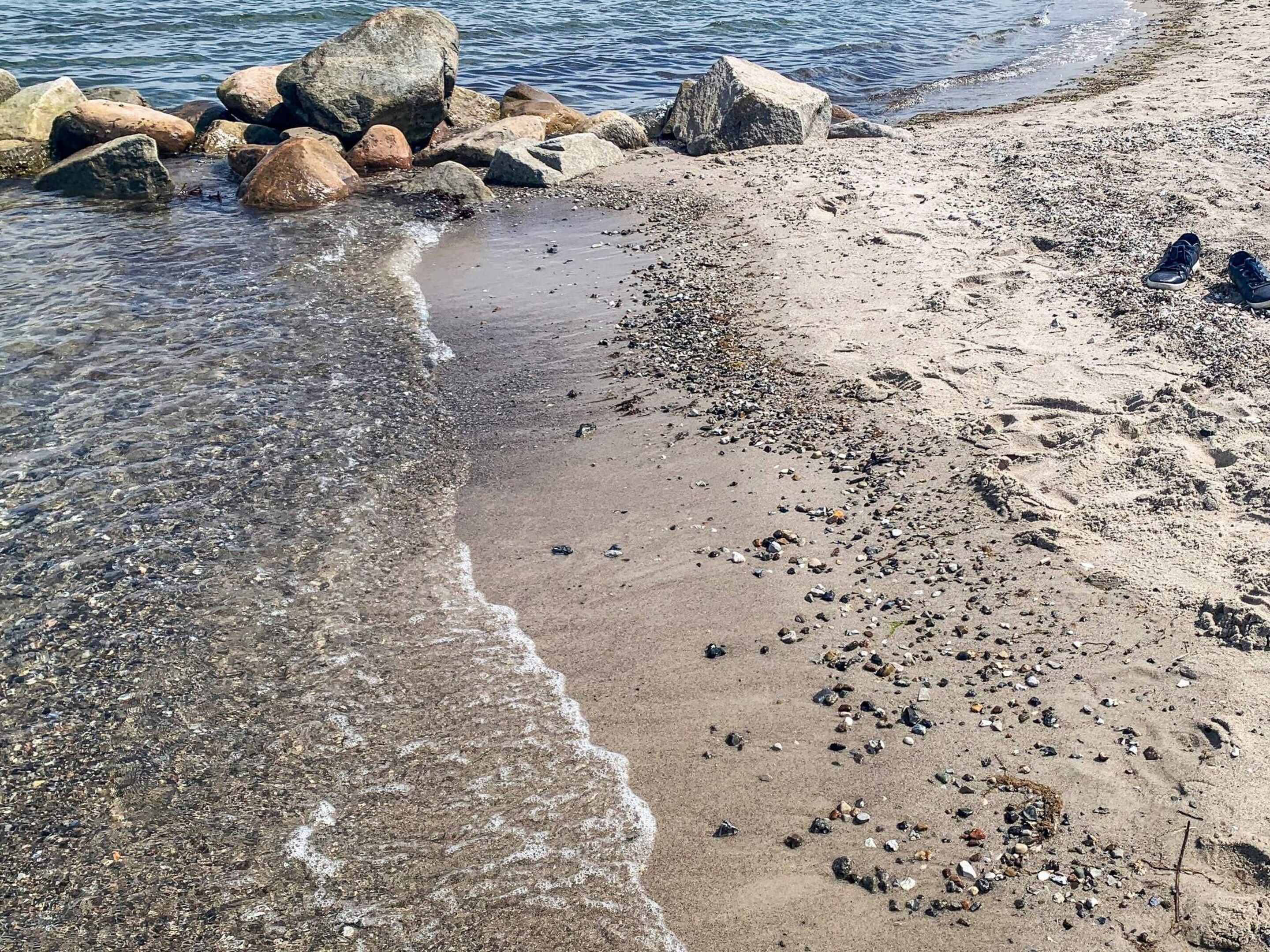 A beach with rocks and pebbles, clear water, and a pair of shoes on the sand.